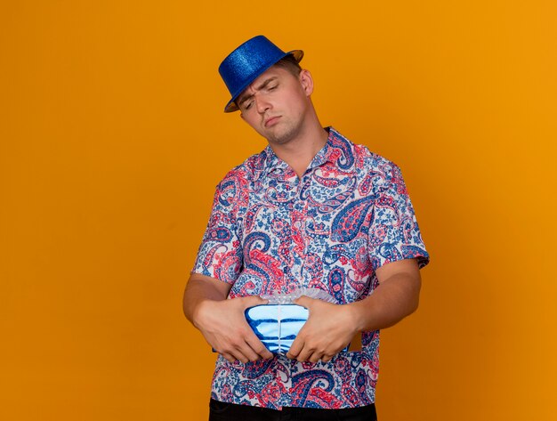 Unzufriedener junger Party-Typ, der blauen Hut hält und Geschenkbox lokalisiert auf Orange hält