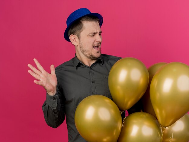 Unzufriedener junger Party-Typ, der blauen Hut hält und Luftballons isoliert auf Rosa hält