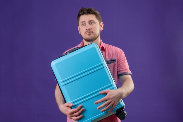 Unzufriedener junger Reisender Mann, der blauen Koffer hält, der mit traurigem Ausdruck beiseite schaut