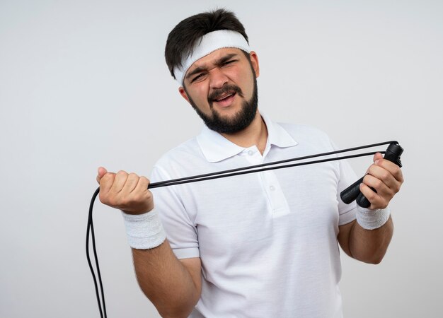 Unzufriedener junger sportlicher Mann, der Stirnband und Armband trägt, die ein Springseil strecken, das auf weißer Wand isoliert wird