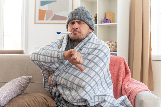 Unzufriedener kranker Mann mit Schal um den Hals, der eine in Plaid gehüllte Wintermütze trägt, die seine Temperatur mit einem Thermometer misst und auf der Couch im Wohnzimmer sitzt