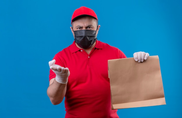 Unzufriedener Lieferbote, der rote Uniform und Kappe in der Gesichtsschutzmaske trägt, die Papierpaket hält, das mit Arm der Hand zur Kamera zeigt, die über blauem Raum stehend argumentiert