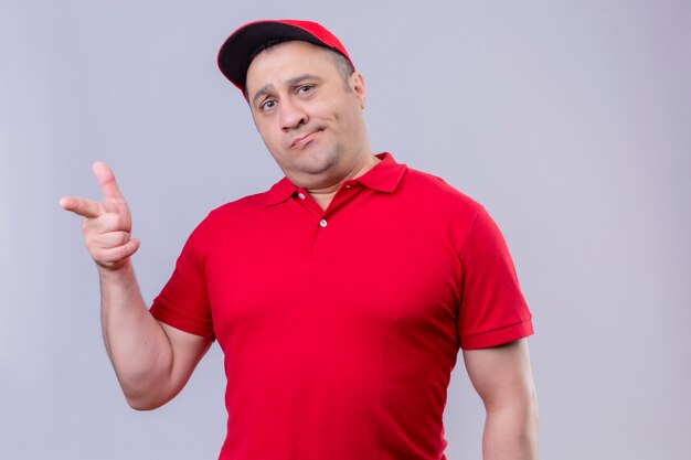 Unzufriedener Lieferbote in roter Uniform und Mütze mit skeptischem Gesichtsausdruck, der mit dem Zeigefinger auf die Kamera über der weißen Wand zeigt