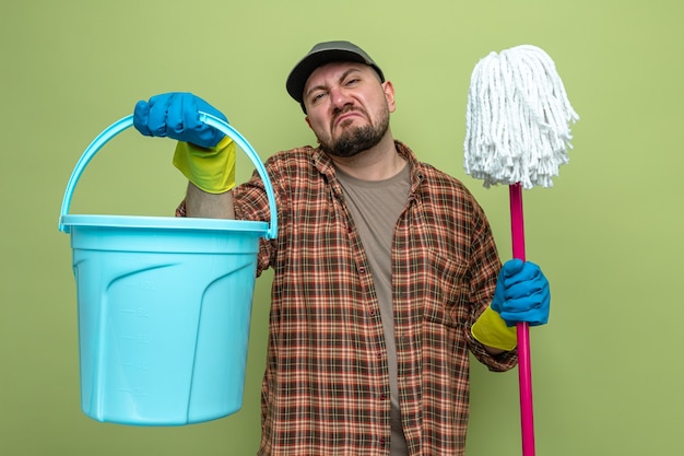 Kostenloses Foto unzufriedener slawischer putzmann mit gummihandschuhen, der eimer und mopp hält