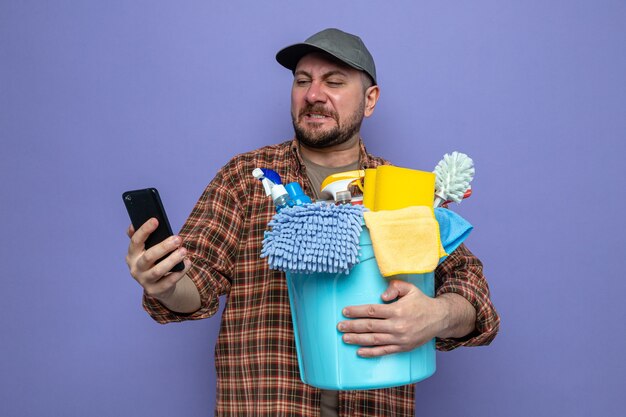 Unzufriedener slawischer Reinigungsmann, der Reinigungsgeräte hält und auf das Telefon schaut