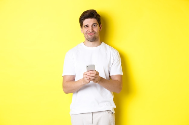 Unzufriedener und widerstrebender Mann, der eine Grimasse verzieht, von der Nachricht auf dem Smartphone nicht amüsiert ist und über gelbem Hintergrund steht