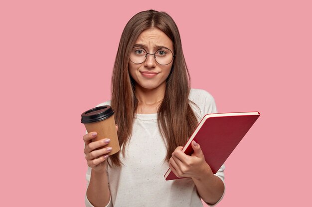 Unzufriedener unglücklicher Student sieht missfallen aus, runzelt die Stirn, trägt eine optische Brille, trägt ein Lehrbuch und ein heißes Getränk, isoliert über einer rosa Wand, will nicht lernen. Prüfungsvorbereitung.
