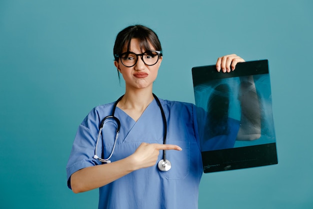 Unzufriedenes Halten und Punkte auf Röntgenbild junge Ärztin mit einheitlichem Fith-Stethoskop isoliert auf blauem Hintergrund