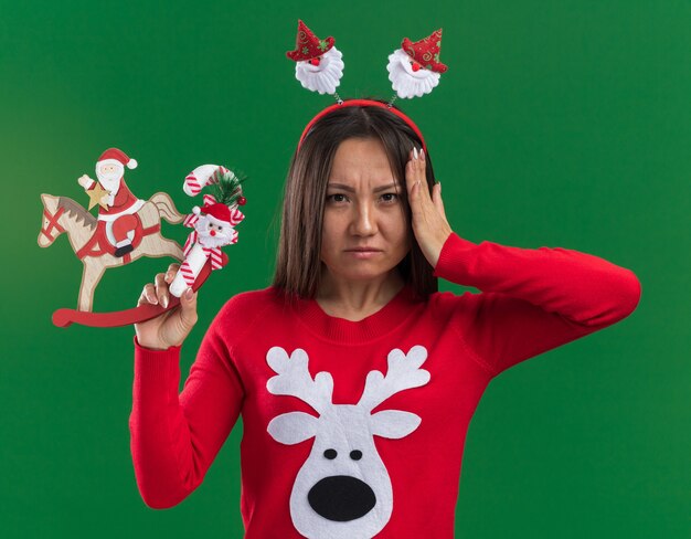 Unzufriedenes junges asiatisches Mädchen, das Weihnachtshaarbügel mit Pullover hält, der Weihnachtsspielzeug mit Süßigkeiten setzt Hand auf Kopf lokalisiert auf grünem Hintergrund hält