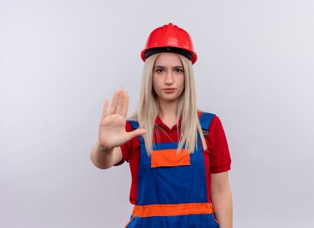 Unzufriedenes junges blondes Ingenieur-Baumeistermädchen in der Uniform, die Hand ausstreckt, die nicht auf isolierte weiße Wand mit Kopienraum gestikuliert