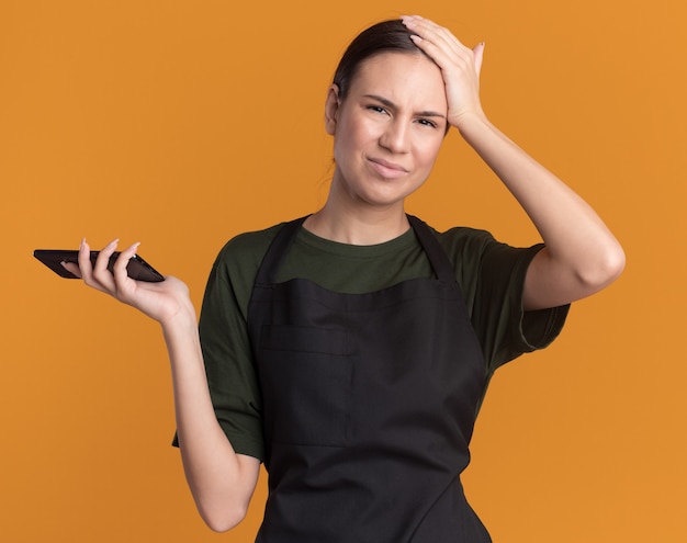 Unzufriedenes junges brünettes Friseurmädchen in Uniform legt die Hand auf die Stirn und hält eine Haarschneidemaschine