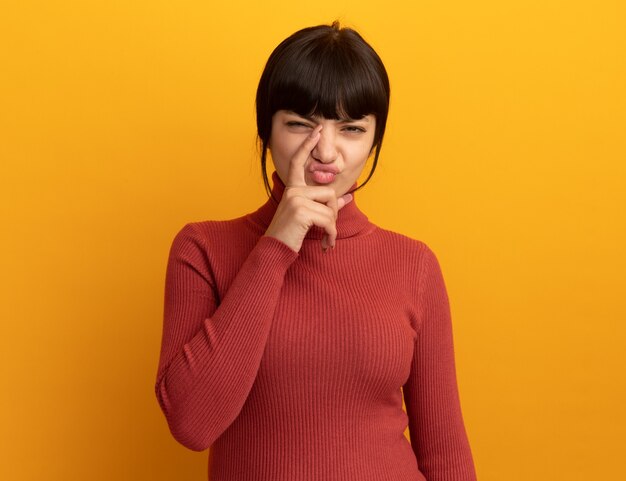 Unzufriedenes junges brünettes kaukasisches Mädchen legt Finger auf die Nase, die auf orangefarbener Wand mit Kopienraum isoliert ist