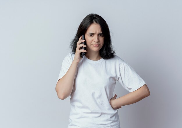 Unzufriedenes junges hübsches kaukasisches Mädchen, das Hand auf Taille setzt, gerade schaut und am Telefon lokalisiert auf weißem Hintergrund mit Kopienraum spricht