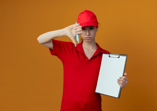 Kostenloses Foto unzufriedenes junges hübsches liefermädchen in der roten uniform und in der kappe, die stift und zwischenablage hält, zeigt daumen unten lokalisiert auf orange hintergrund mit kopienraum