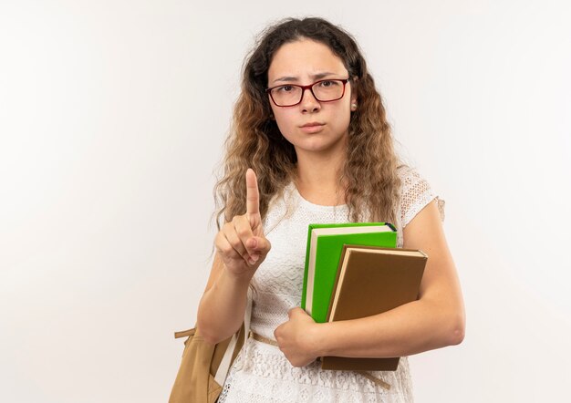 Unzufriedenes junges hübsches Schulmädchen, das Brille und Rückentasche hält Bücher hält Finger hebt lokalisiert auf weißem Hintergrund mit Kopienraum