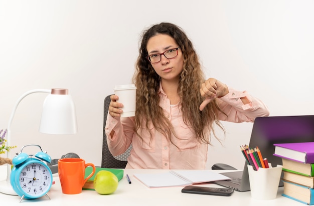 Unzufriedenes junges hübsches Schulmädchen, das eine Brille trägt, die am Schreibtisch mit Schulwerkzeugen sitzt, die ihre Hausaufgaben halten Kaffeetasse halten und Daumen unten lokalisiert auf weißem Hintergrund zeigen