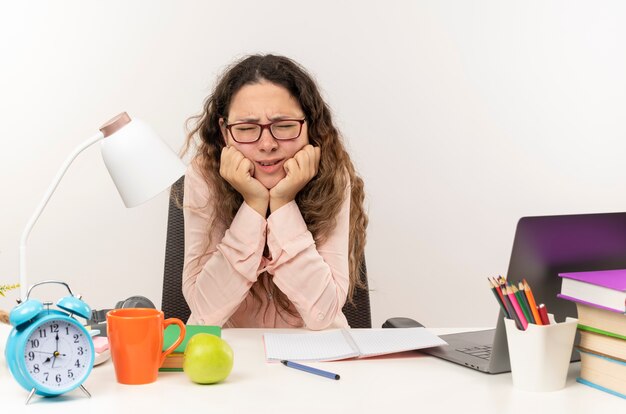 Unzufriedenes junges hübsches Schulmädchen, das eine Brille trägt, die am Schreibtisch mit Schulwerkzeugen sitzt und ihre Hausaufgaben macht, die Hände auf Kinn mit geschlossenen Augen lokalisiert auf weißem Hintergrund setzen