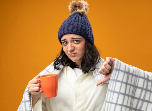 Unzufriedenes junges kaukasisches krankes Mädchen, das Robe Wintermütze in Plaid hält Tasse Tasse Tee mit Blick auf Kamera zeigt Daumen unten isoliert auf orange Hintergrund