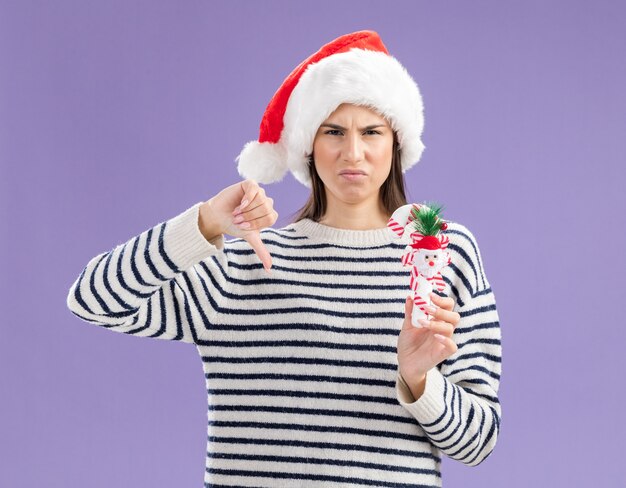 Unzufriedenes junges kaukasisches Mädchen mit Weihnachtsmütze hält Zuckerstange und Daumen nach unten