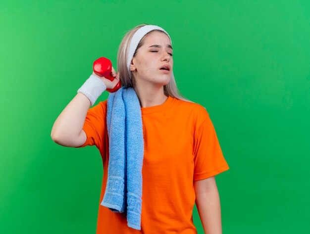 Unzufriedenes junges kaukasisches sportliches Mädchen mit Hosenträgern, das Stirnband und Armbänder mit Handtuch auf der Schulter trägt, hält Hantel du