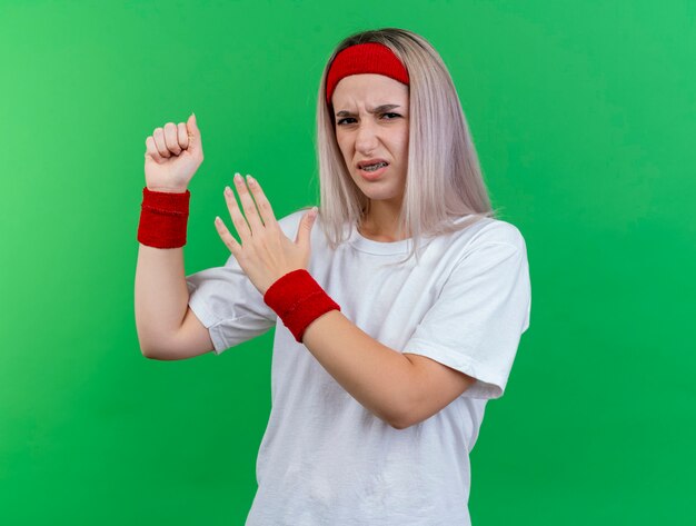 Unzufriedenes junges kaukasisches sportliches Mädchen mit Hosenträgern, das Stirnband und Armbänder trägt, hält die Faust und hält die Hand offen