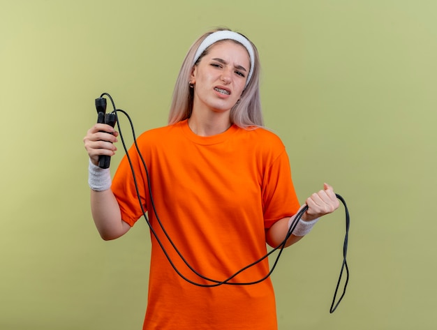 Unzufriedenes junges kaukasisches sportliches Mädchen mit Hosenträgern, das Stirnband und Armbänder trägt, hält Springseil