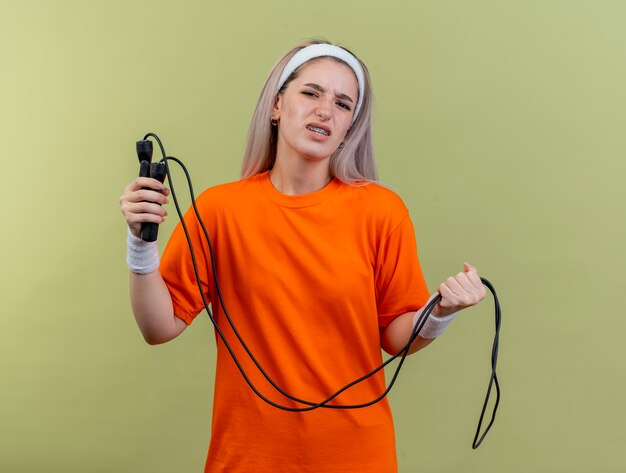Unzufriedenes junges kaukasisches sportliches Mädchen mit Hosenträgern, das Stirnband und Armbänder trägt, hält Springseil