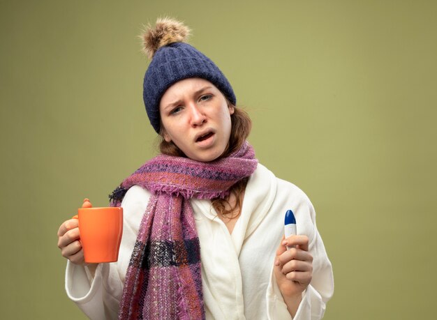 Unzufriedenes junges krankes Mädchen, das weiße Robe und Wintermütze mit Schal hält Tasse Tee mit Thermometer auf Olivgrün mit Kopienraum isoliert