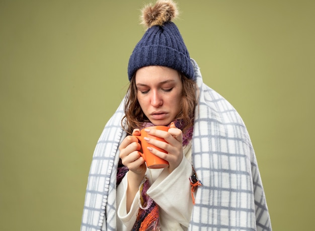 Kostenloses Foto unzufriedenes junges krankes mädchen, das weiße robe und wintermütze mit schal wickelt, der im karierten halten und betrachten der tasse tee lokalisiert auf olivgrün eingewickelt ist