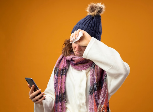 Unzufriedenes junges krankes Mädchen, das weißes Gewand und Wintermütze mit Schal hält und Telefon betrachtet, das Hand auf Stirn lokalisiert auf orange Wand setzt