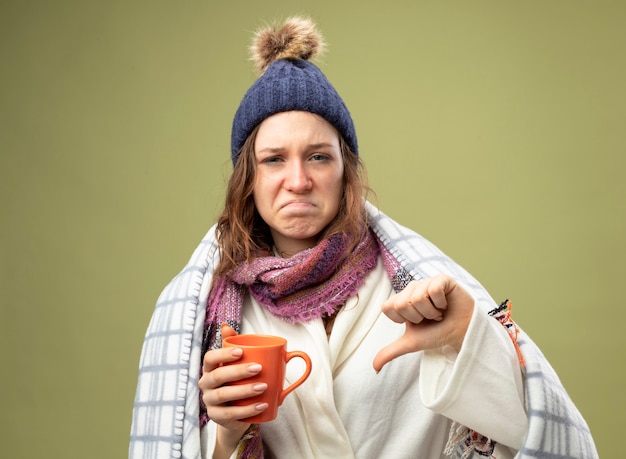 Unzufriedenes junges krankes Mädchen, das weißes Gewand und Wintermütze mit Schal wickelt in Plaid hält Tasse Tee zeigt Daumen nach unten isoliert auf Olivgrün