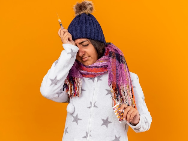 Unzufriedenes junges krankes Mädchen mit geschlossenen Augen, das Wintermütze mit Schal hält, der Pillen mit Spritze hält und Hand auf Stirn lokalisiert auf gelbem Hintergrund setzt