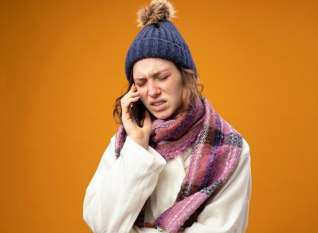 Unzufriedenes junges krankes Mädchen mit geschlossenen Augen, die weiße Robe und Wintermütze mit Schal tragen, spricht am Telefon lokalisiert auf orange Wand