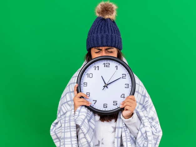 Unzufriedenes junges krankes Mädchen mit geschlossenen Augen, die Wintermütze mit Schal bedeckten Gesicht mit Wanduhr lokalisiert auf grünem Hintergrund tragen