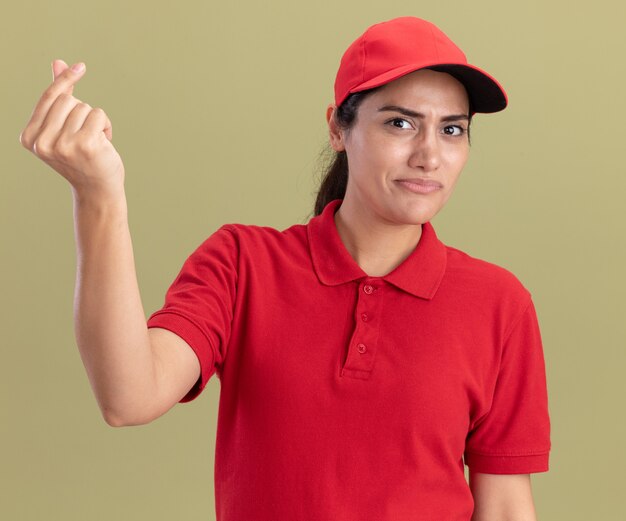 Unzufriedenes junges Liefermädchen, das Uniform mit Kappe trägt, die Spitzengeste zeigt, die auf olivgrüner Wand lokalisiert wird