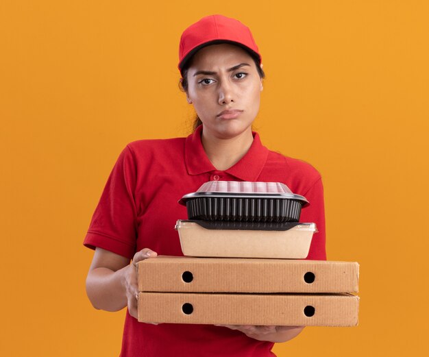 Unzufriedenes junges Liefermädchen in Uniform und Mütze mit Pizzakartons mit Lebensmittelbehältern isoliert auf oranger Wand