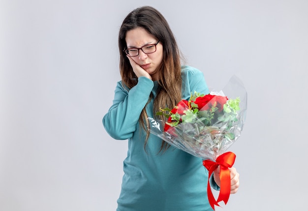 Unzufriedenes junges Mädchen am Valentinstag, das Blumenstrauß hält und anschaut und die Hand auf die Wange legt, isoliert auf weißem Hintergrund