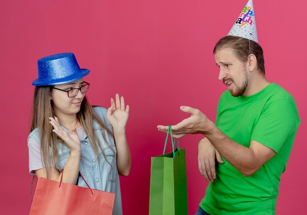 Unzufriedenes junges Mädchen, das blauen Partyhut trägt, hält rote Geschenktüte und hebt Hände, die nicht auf unzuverlässigen jungen Mann schauend zeigen, der Partyhut trägt und grüne Geschenktüte lokalisiert auf rosa Wand hält