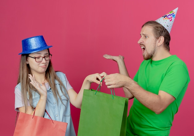 Kostenloses Foto unzufriedenes junges mädchen, das blauen partyhut trägt, hält rote geschenktüte und hebt hand, die grüne geschenktüte vom verärgerten jungen mann trägt, der partyhut isoliert auf rosa wand trägt