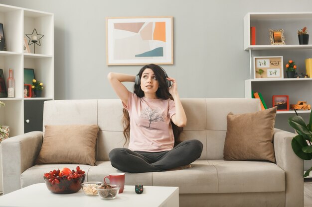 Unzufriedenes junges Mädchen mit Kopfhörern, das auf dem Sofa hinter dem Couchtisch im Wohnzimmer sitzt