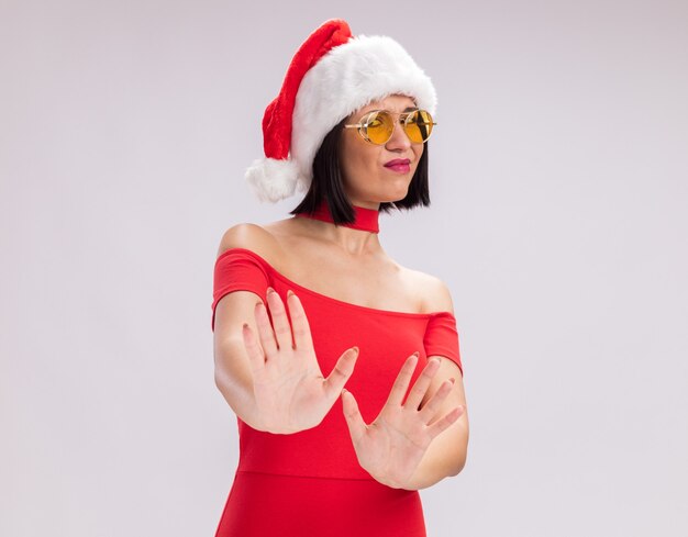 Unzufriedenes junges Mädchen mit Weihnachtsmütze und Brille, das in die Kamera schaut und die Ablehnungsgeste isoliert auf weißem Hintergrund mit Kopierraum macht
