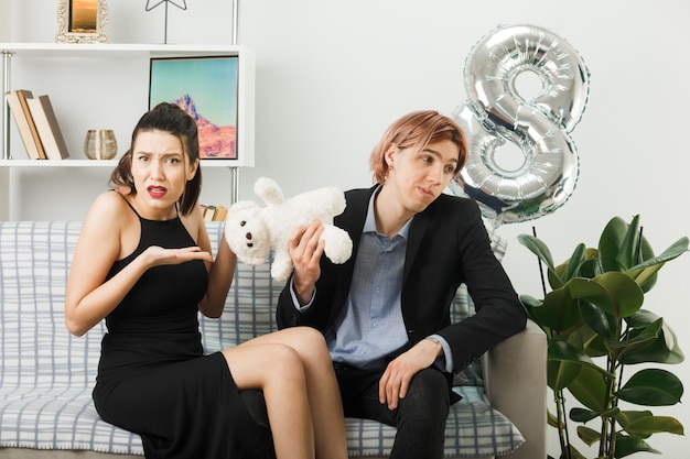 Unzufriedenes junges Paar am glücklichen Frauentag mit Teddybär, der auf dem Sofa im Wohnzimmer sitzt