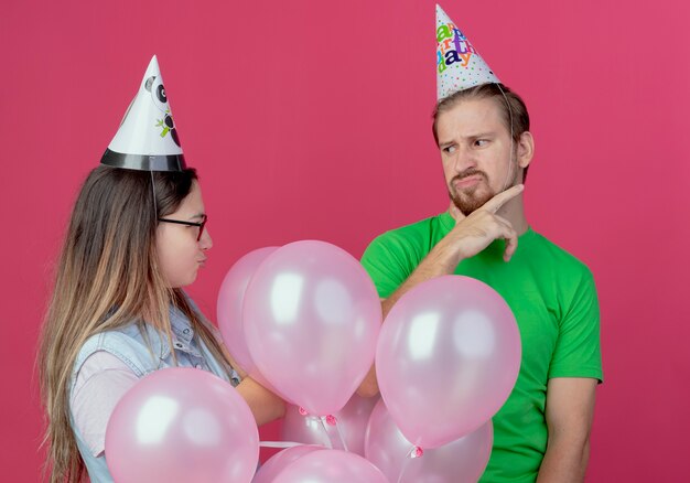 Unzufriedenes junges Paar, das Partyhut trägt, schaut einander stehend mit Heliumballons an, die auf rosa Wand isoliert werden
