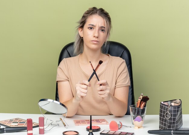 Unzufriedenes junges schönes Mädchen, das am Schreibtisch mit Make-up-Tools sitzt und Make-up-Pinsel auf olivgrünem Hintergrund hält und kreuzt