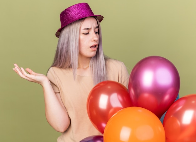 Unzufriedenes junges schönes Mädchen mit Partyhut, das hinter Ballons steht, die Hand isoliert auf olivgrüner Wand ausbreitet