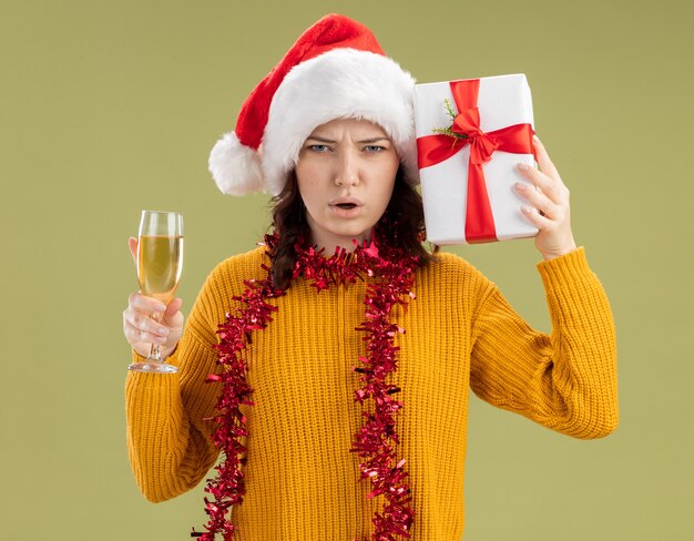 Unzufriedenes junges slawisches Mädchen mit Weihnachtsmütze und mit Girlande um Hals, die Glas Champagner und Weihnachtsgeschenkbox lokalisiert auf olivgrünem Hintergrund mit Kopienraum hält