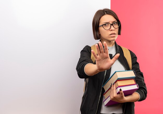 Unzufriedenes junges Studentenmädchen, das Brille und Rückentasche hält Bücher hält, die vor weißer Wand stehen, die nicht lokalisiert auf rosa Hintergrund mit Kopienraum gestikuliert