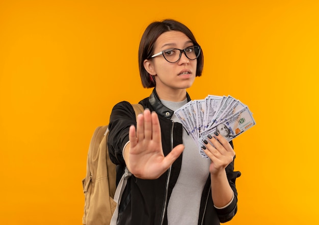 Unzufriedenes junges Studentenmädchen, das Brille und Rückentasche hält, die Geld gestikulieren Stopp lokalisiert auf orange Hintergrund mit Kopienraum