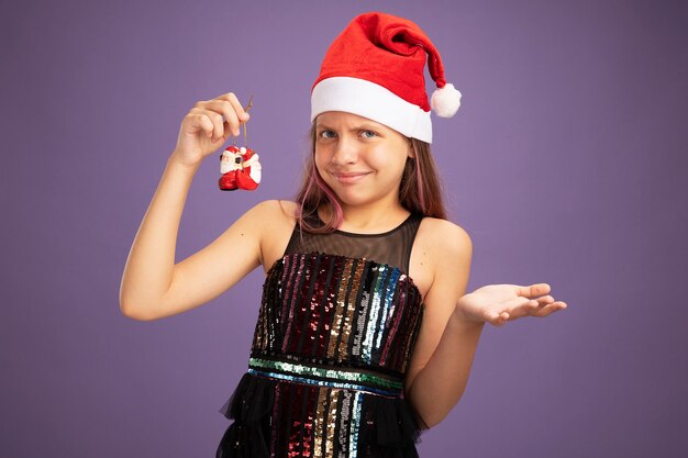 Unzufriedenes kleines Mädchen in Glitzer-Partykleid und Weihnachtsmütze mit Weihnachtsspielzeug, das in die Kamera schaut und die Hand in Unmut und Empörung über violettem Hintergrund hebt