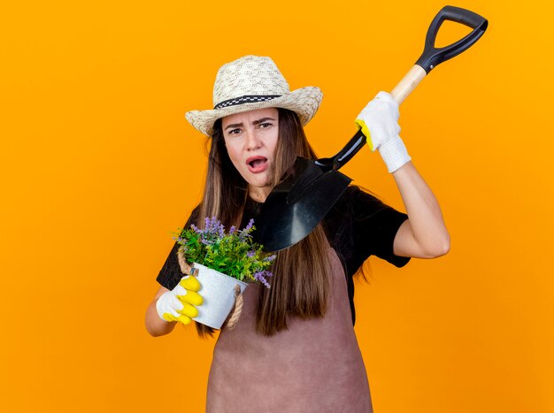 Unzufriedenes schönes Gärtnermädchen, das Uniform und Gartenhut mit Handschuhen hält und Punkte mit Spaten an Blume im Blumentopf lokalisiert auf orangefarbenem Hintergrund trägt